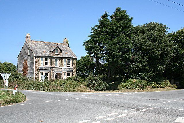 A stone house with a gabled roof, a road intersection, and dense trees in the background.