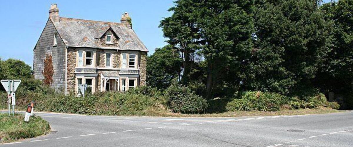 A stone house with a gabled roof, a road intersection, and dense trees in the background.