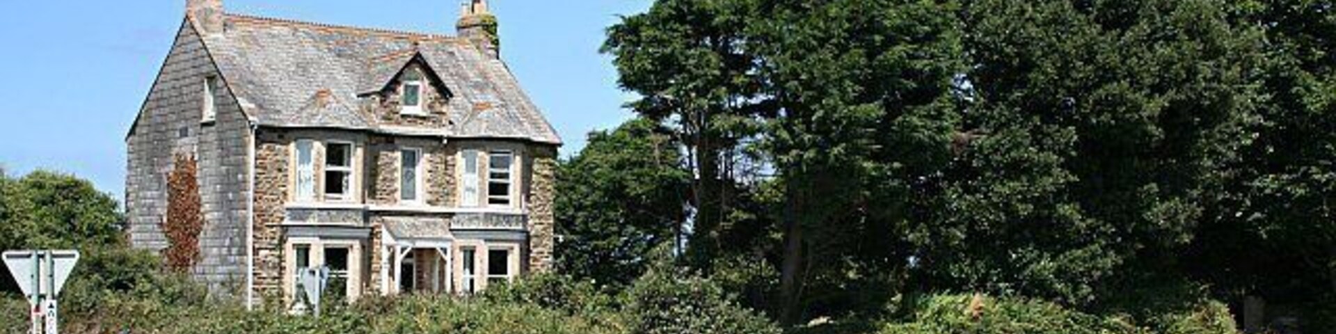 A stone house with a gabled roof, a road intersection, and dense trees in the background.