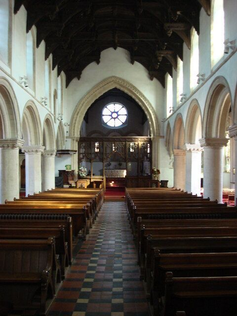 Interior de igreja com bancos de madeira, piso xadrez e uma grande janela circular no altar.