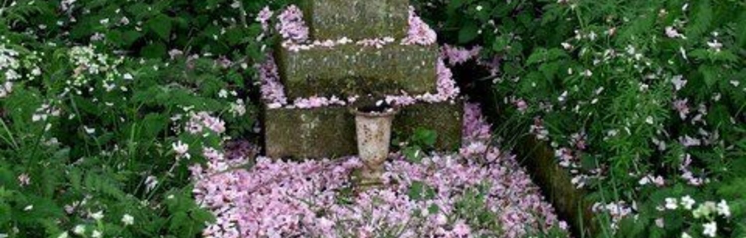 Uma cruz de pedra rodeada por flores cor-de-rosa e vegetação.