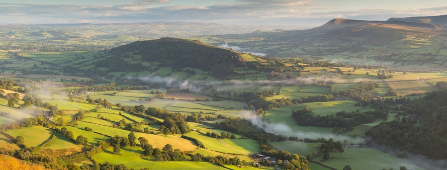 Rolling green hills, a mountain with a smoke plume, and a valley with scattered houses.