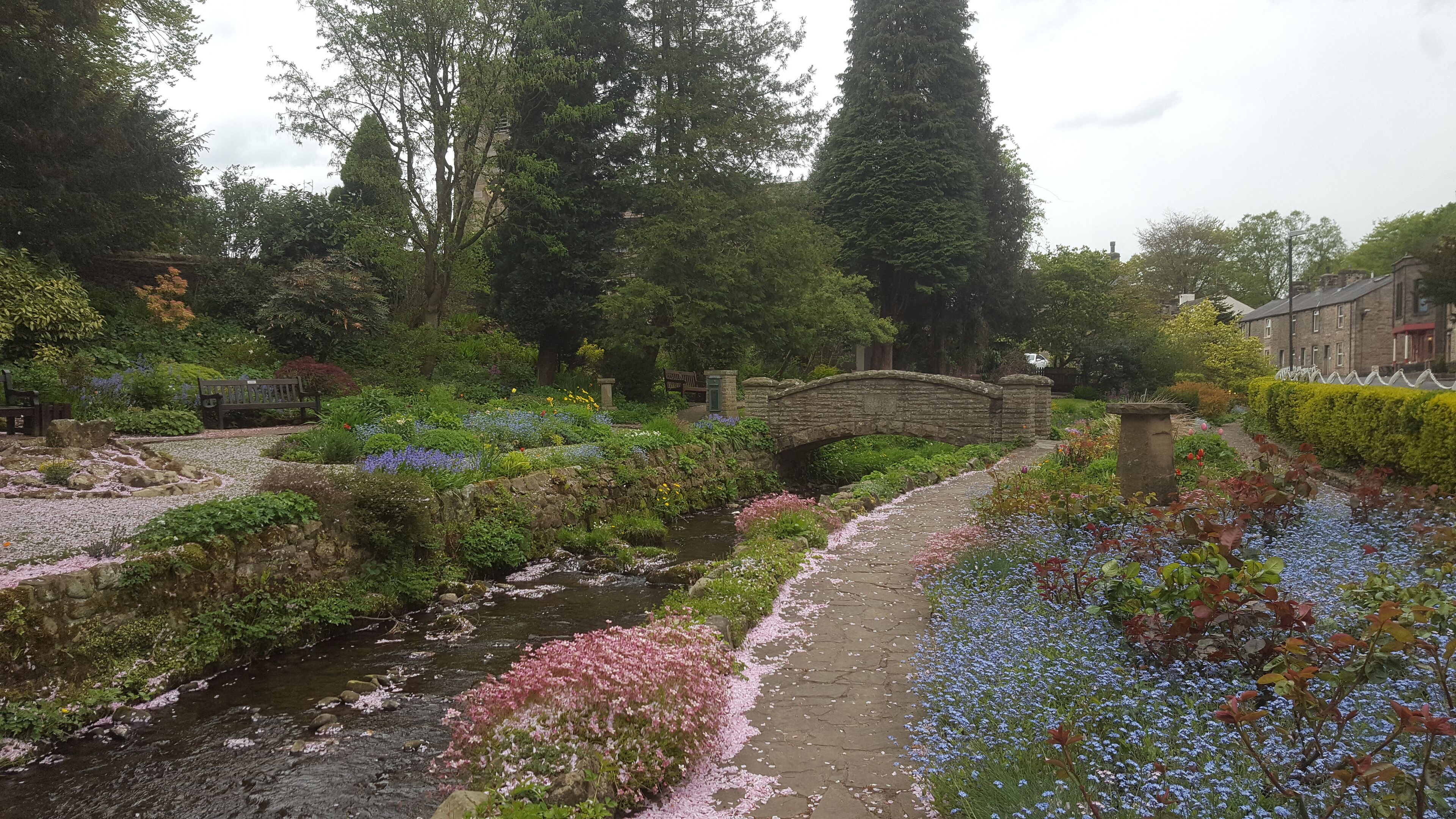 A stone bridge over a stream in a garden with flowering plants and trees.