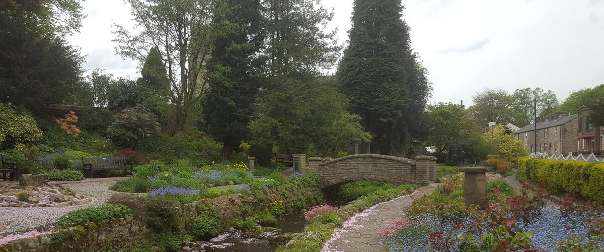 A stone bridge over a stream in a garden with flowering plants and trees.