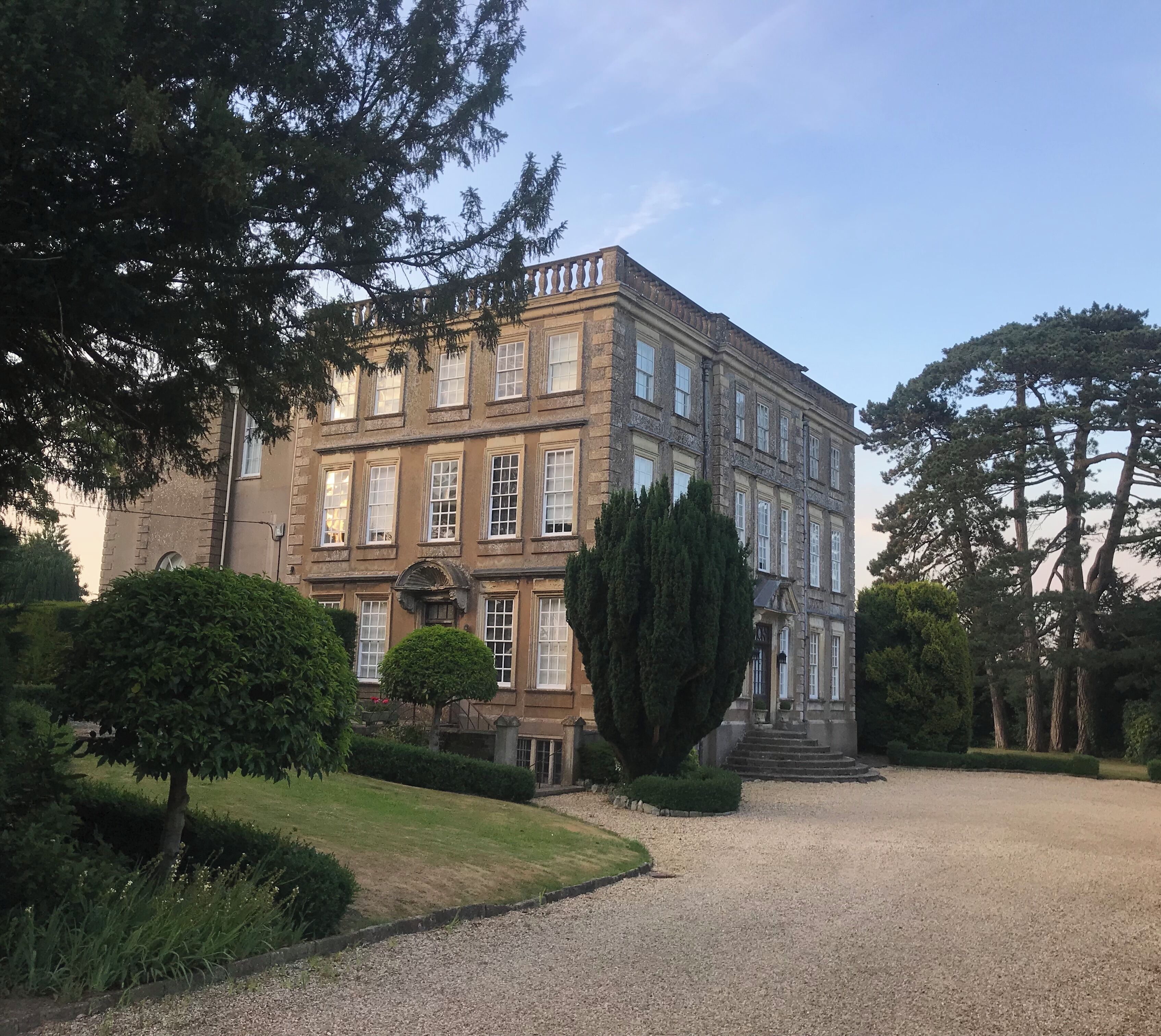 A large, historic building with a symmetrical facade, surrounded by well-maintained gardens and tall trees.