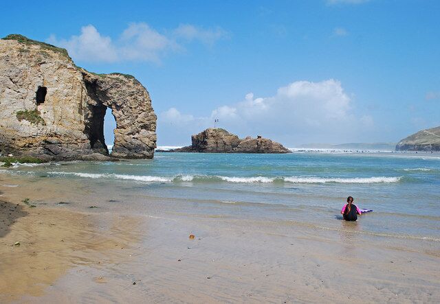 A beach with a large rock formation featuring an arch, waves, and a person sitting on the sand.