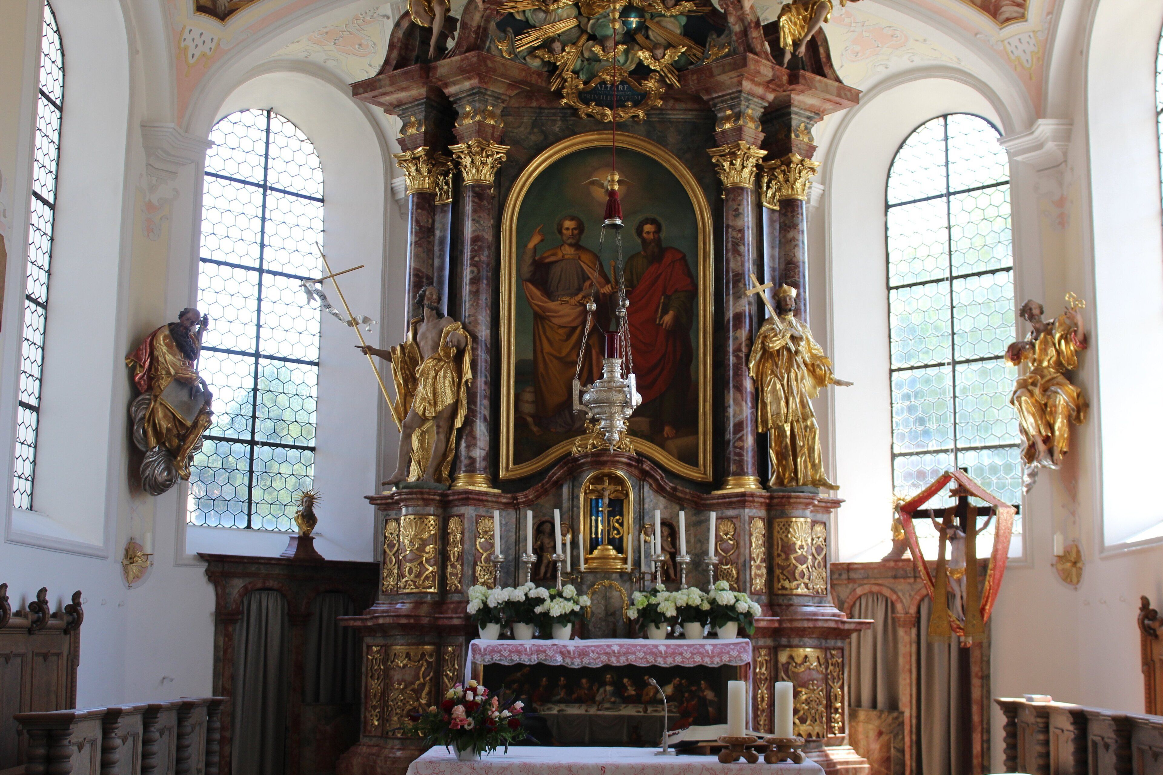 An ornate church interior with a central religious painting, golden statues, and floral arrangements.