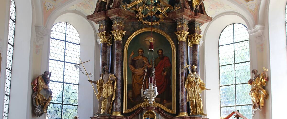 An ornate church interior with a central religious painting, golden statues, and floral arrangements.