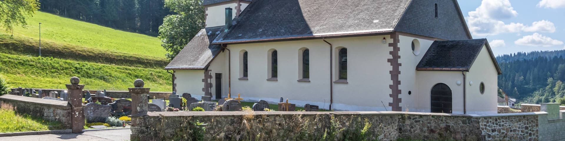 A church with a tall steeple and a cross on top, surrounded by a stone wall and a cemetery.