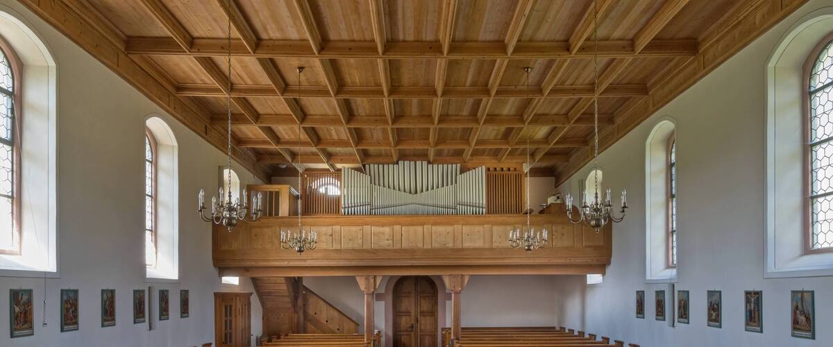 A church interior with wooden pews, a wooden organ loft, and arched windows.