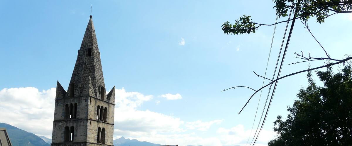 A church with a tall bell tower and a red and white striped roof, surrounded by mountains and greenery.