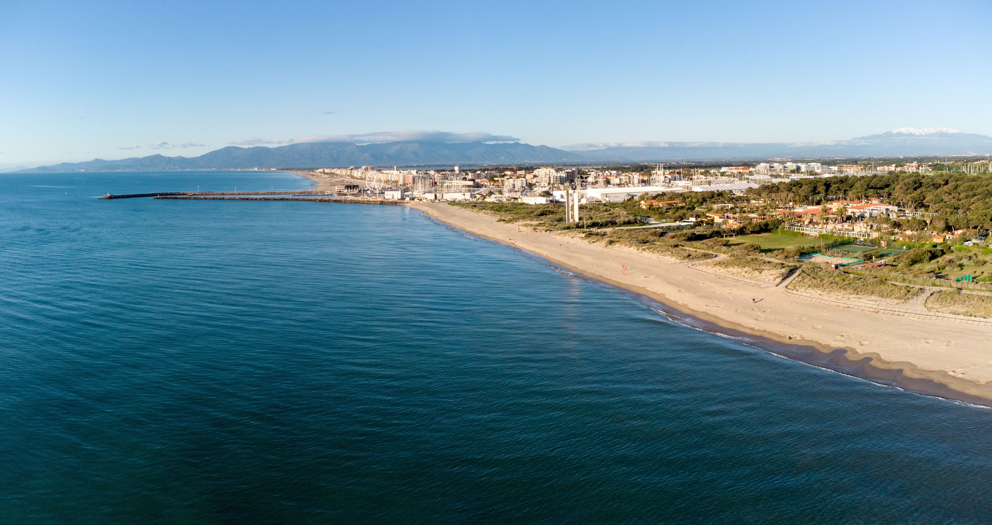 A coastal city with a long beach, a pier extending into the sea, and mountains in the distance.