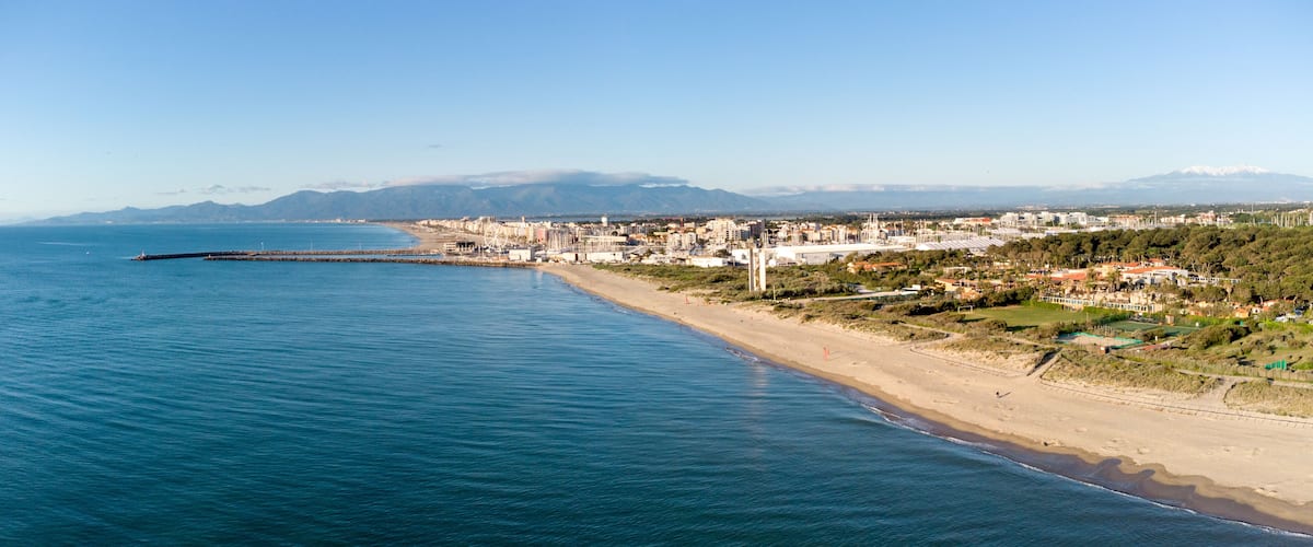 A coastal city with a long beach, a pier extending into the sea, and mountains in the distance.
