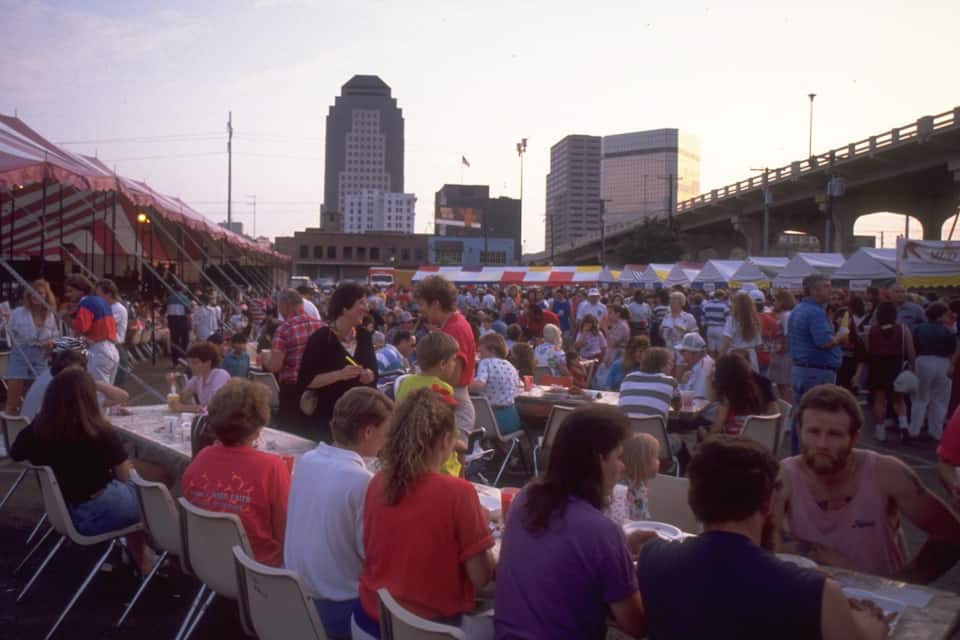 A crowded outdoor event with people seated at tables under tents, with a city skyline and a bridge in the background.