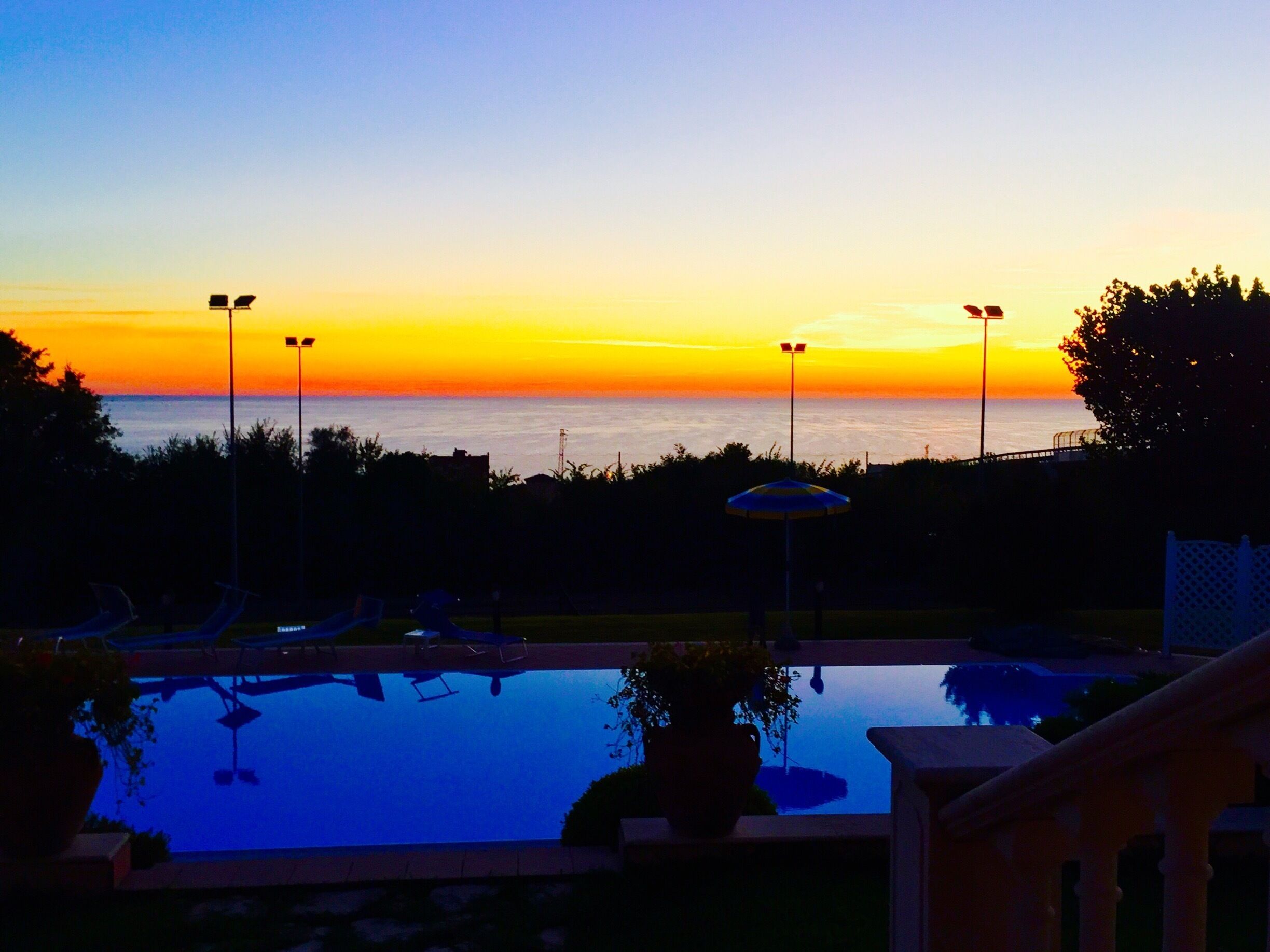 A sunset over a calm pool with lounge chairs and a potted plant.