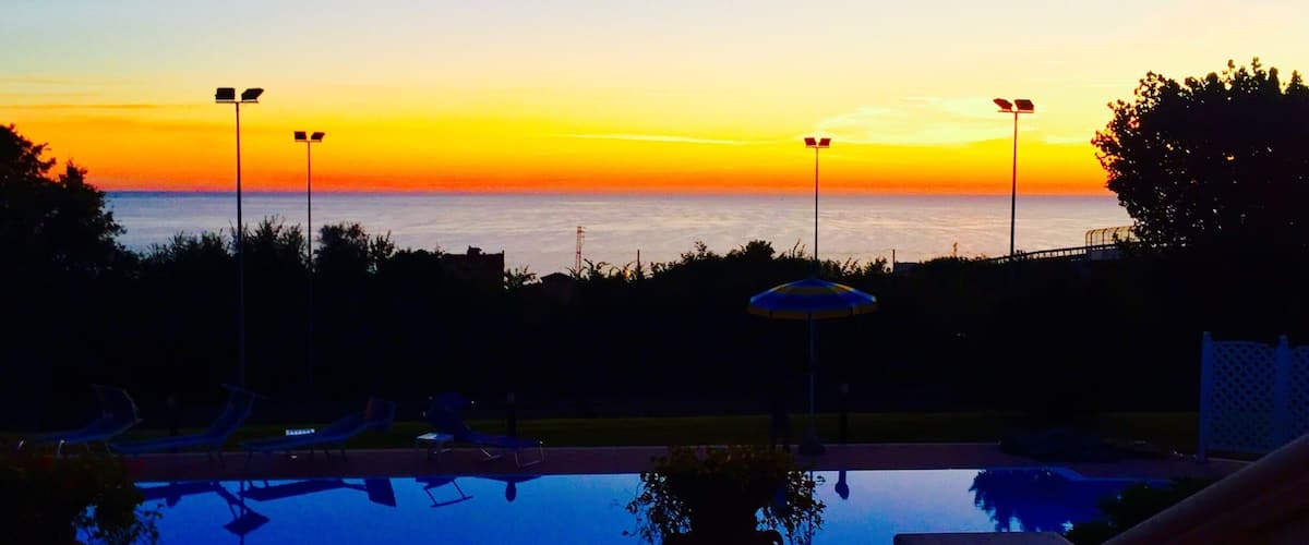 A sunset over a calm pool with lounge chairs and a potted plant.