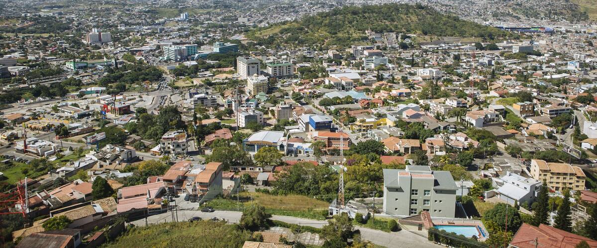A panoramic view of a city with a mix of residential and commercial buildings, surrounded by green hills and mountains in the background.