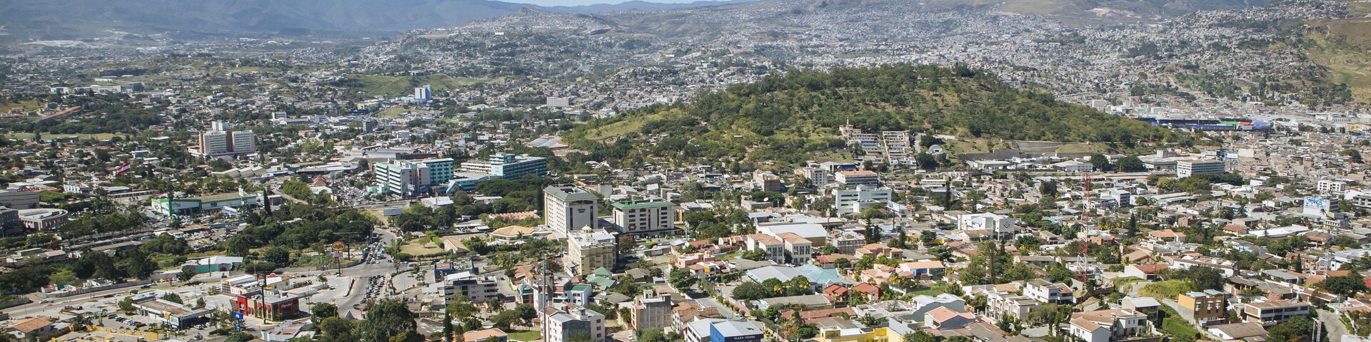 A panoramic view of a city with a mix of residential and commercial buildings, surrounded by green hills and mountains in the background.