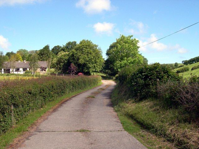 Uma estrada rural estreita ladeada por sebes e árvores, conduzindo a uma casa ao longe.