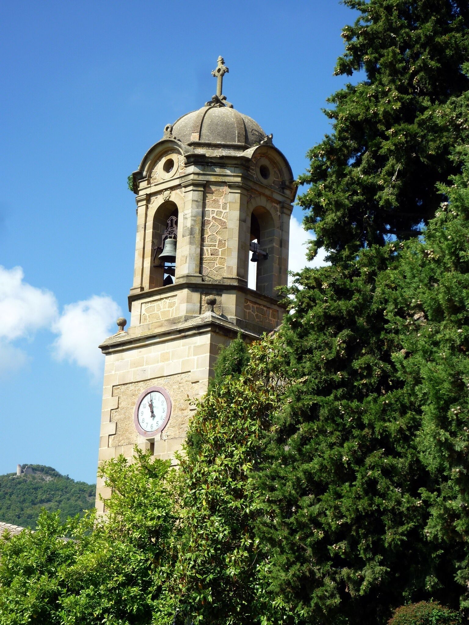 A stone bell tower with a clock, surrounded by trees.