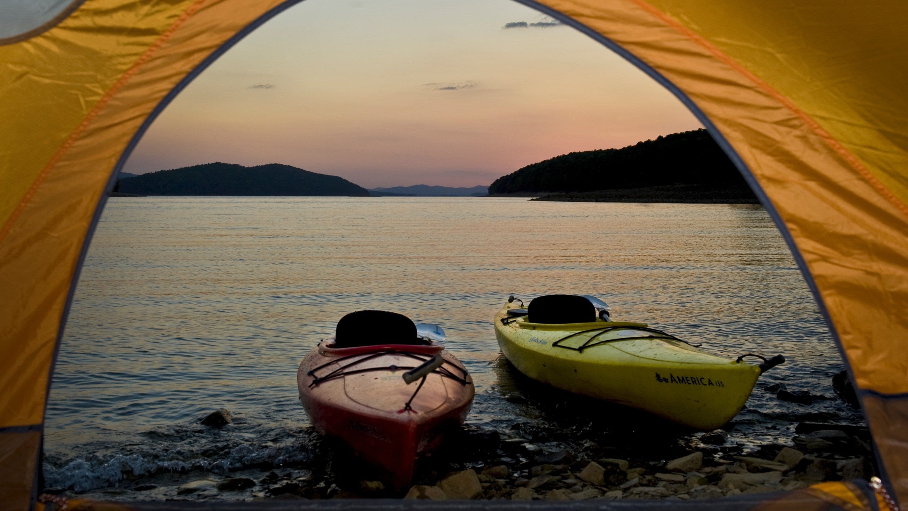 A tent with two kayaks on a rocky shore overlooking a calm body of water at sunset.