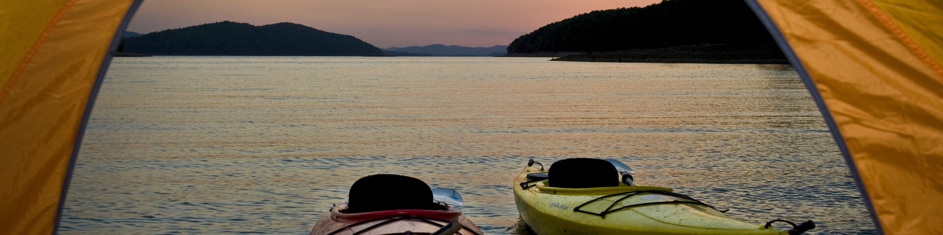 Una carpa con dos kayaks en una orilla rocosa con vistas a una masa de agua tranquila al atardecer.