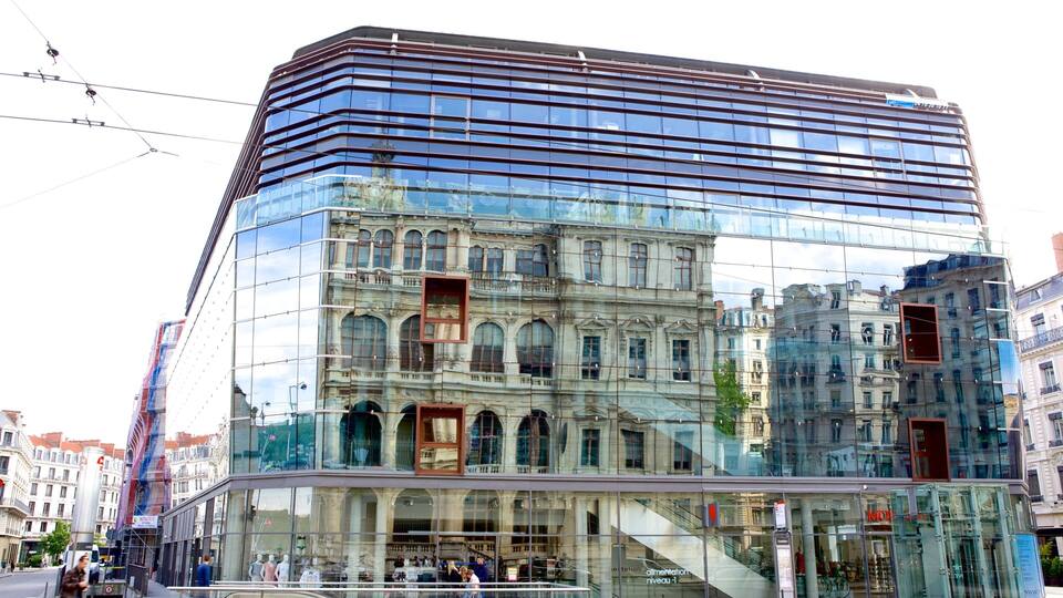 Ein modernes Glasgebäude mit klassischer Fassadenreflexion, eine Straße mit Fußgängern und städtische Umgebung.