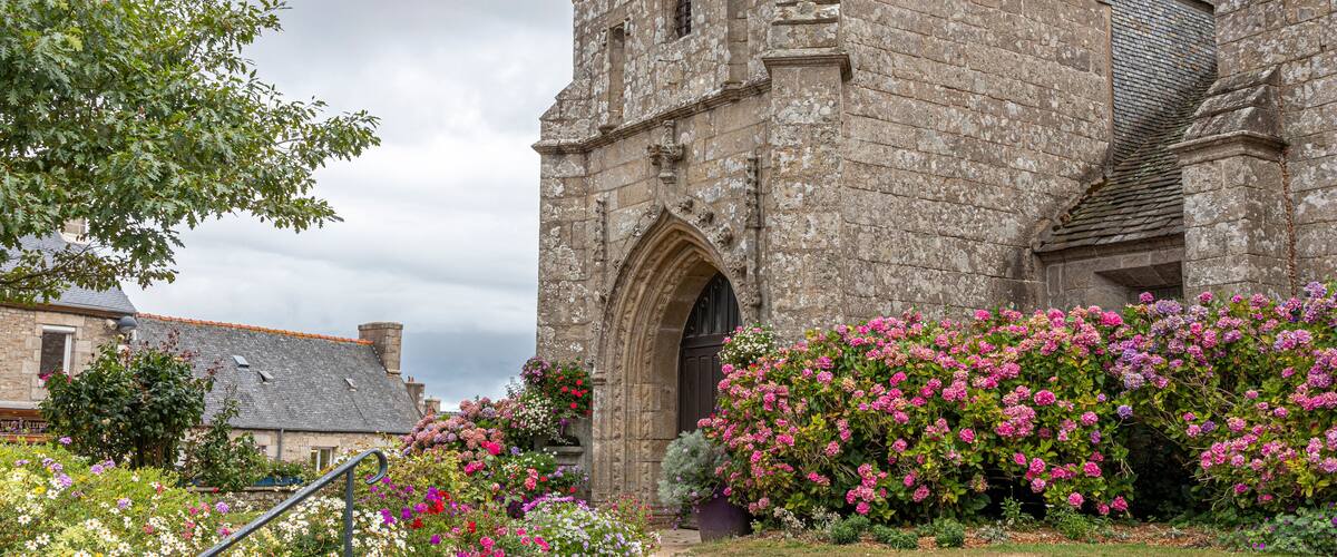 A historic stone building with a clock tower, surrounded by vibrant flowering bushes and a well-maintained garden.