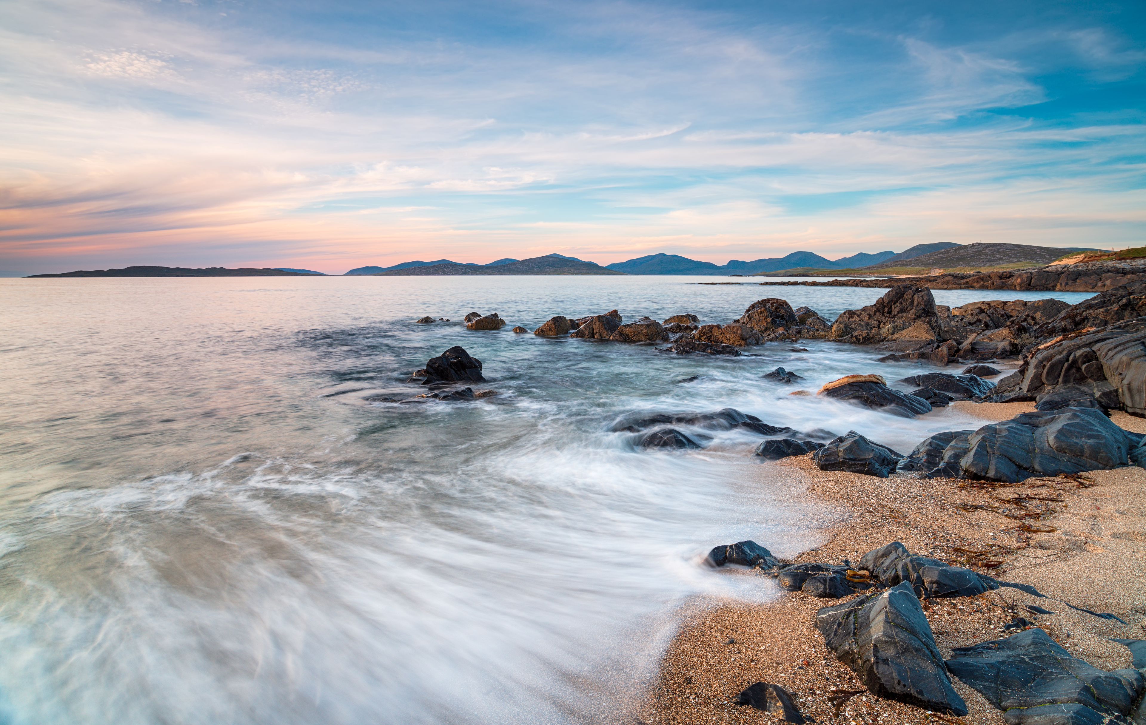 A coastal landscape with rocky shores, calm sea, and distant mountains under a clear sky.