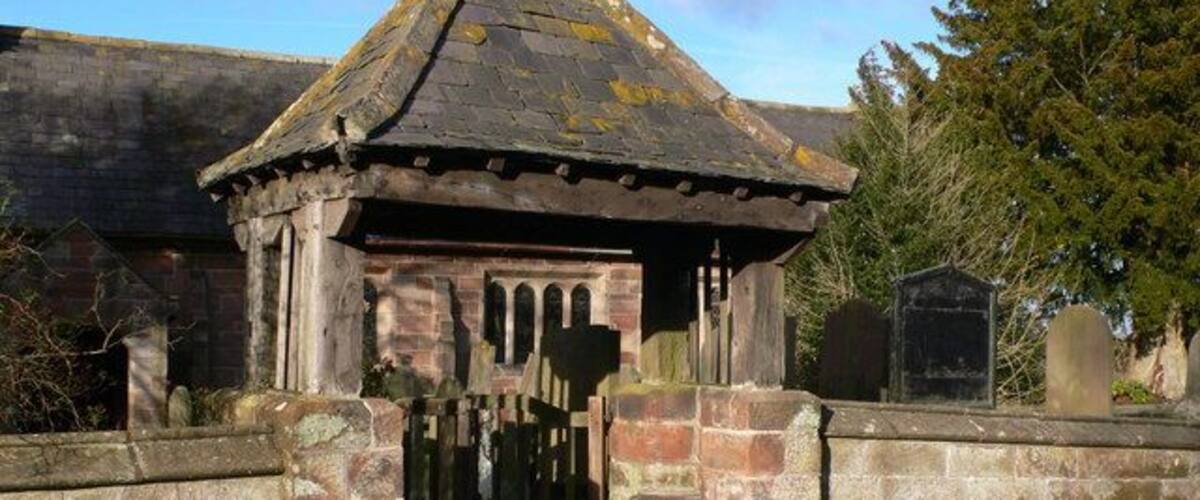 A small stone structure with a tiled roof, surrounded by a low stone wall and a grassy area.