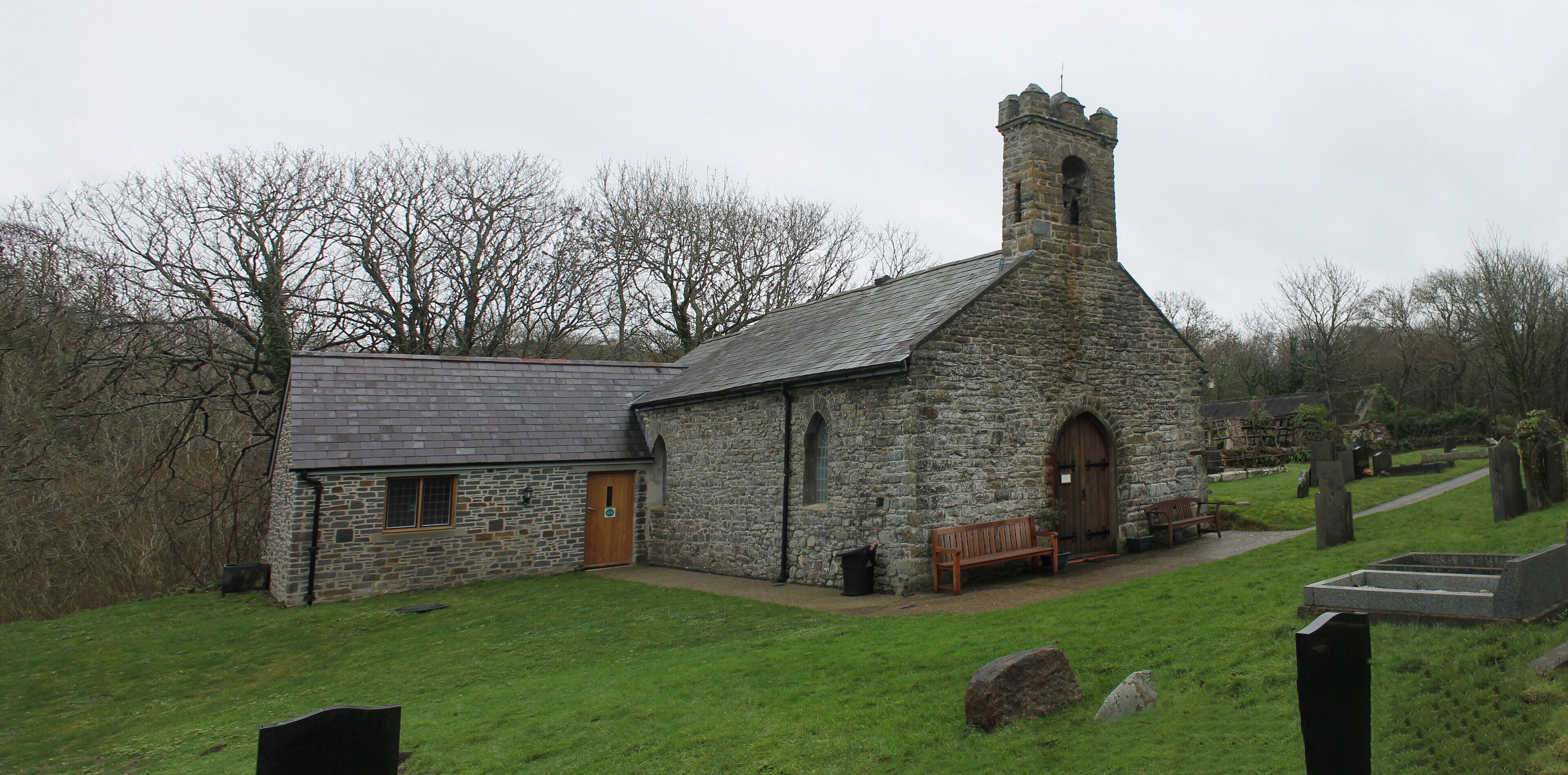 A stone church with a bell tower, a wooden bench, and a cemetery in the background.