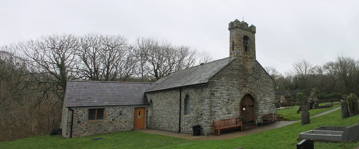 A stone church with a bell tower, a wooden bench, and a cemetery in the background.