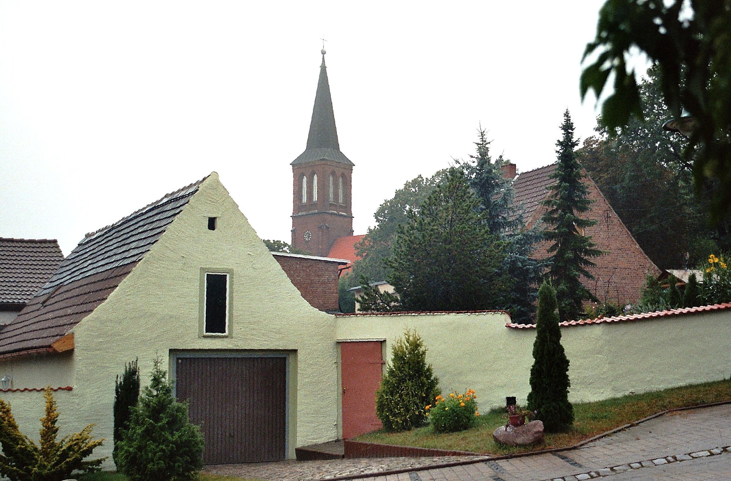 A church with a tall steeple, a small building with a sloped roof, and a paved area with a statue.