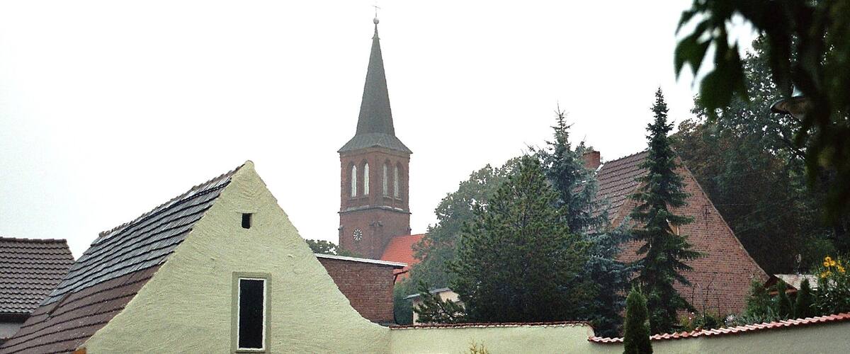 A church with a tall steeple, a small building with a sloped roof, and a paved area with a statue.