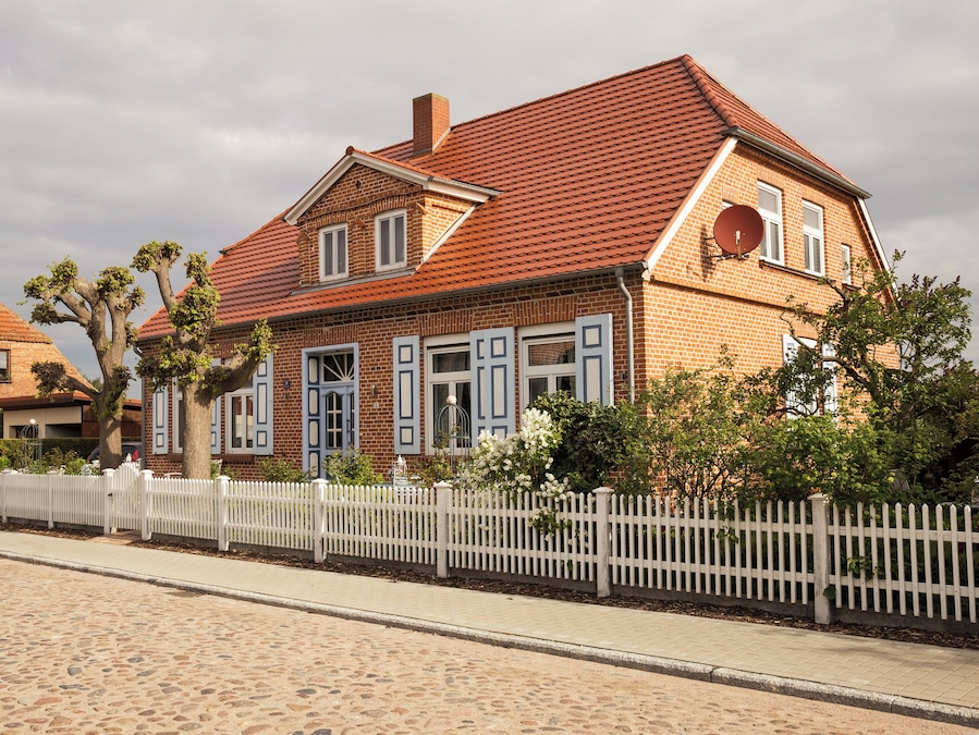 Ein Haus aus Ziegeln mit rotem Ziegeldach, weißen Fensterläden und einem weißen Zäunchen.