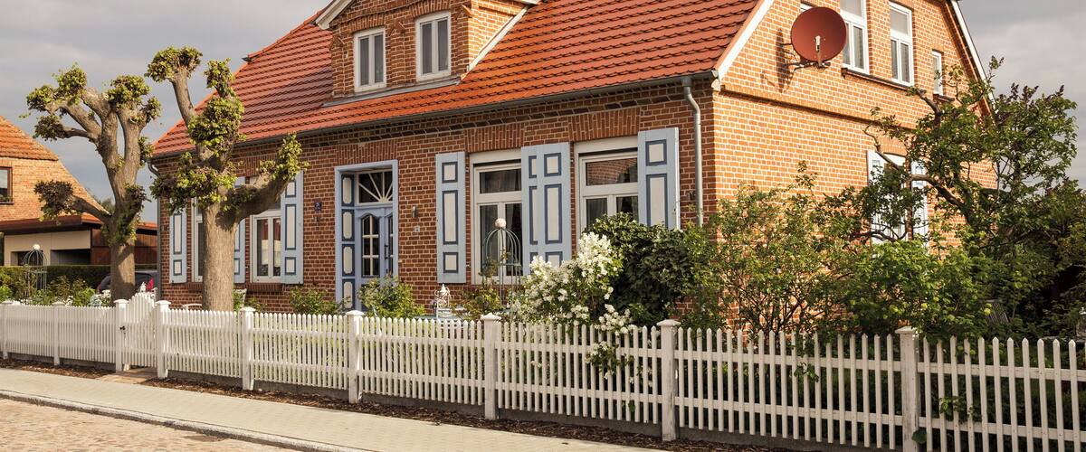 A brick house with a red tiled roof, white shutters, and a white picket fence.