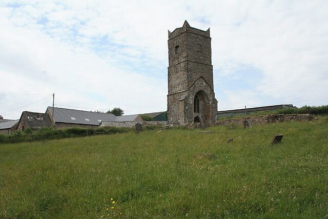 A stone church tower with a bell tower, surrounded by a grassy field and a few buildings in the background.