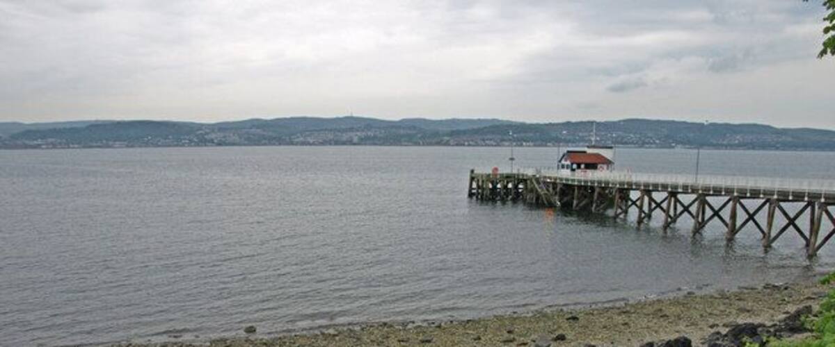 A rocky shoreline with a pier extending into a calm body of water, surrounded by greenery and distant hills.