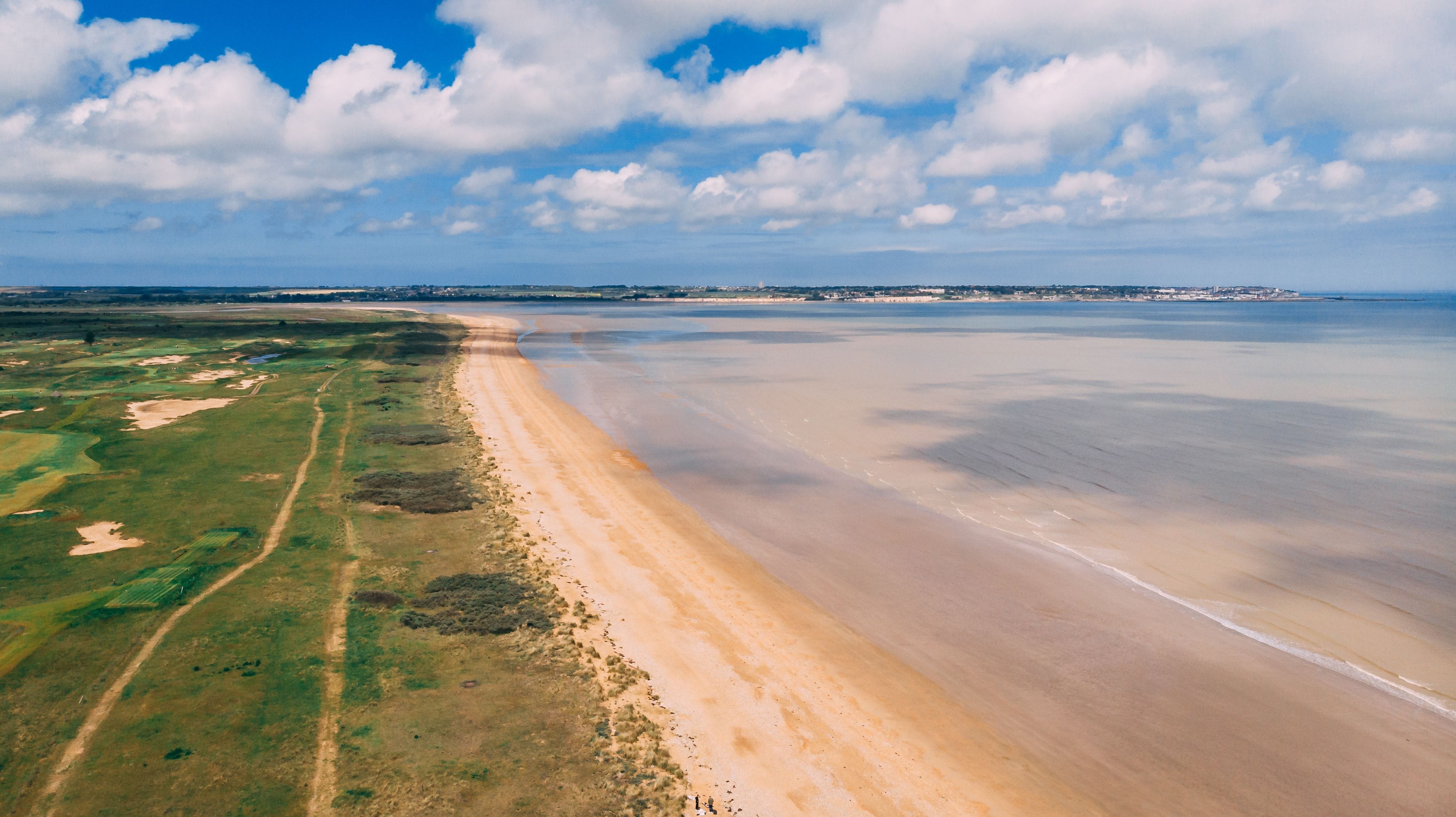 Un paisaje costero con playa arenosa, vegetación verde y cielo azul despejado con nubes.