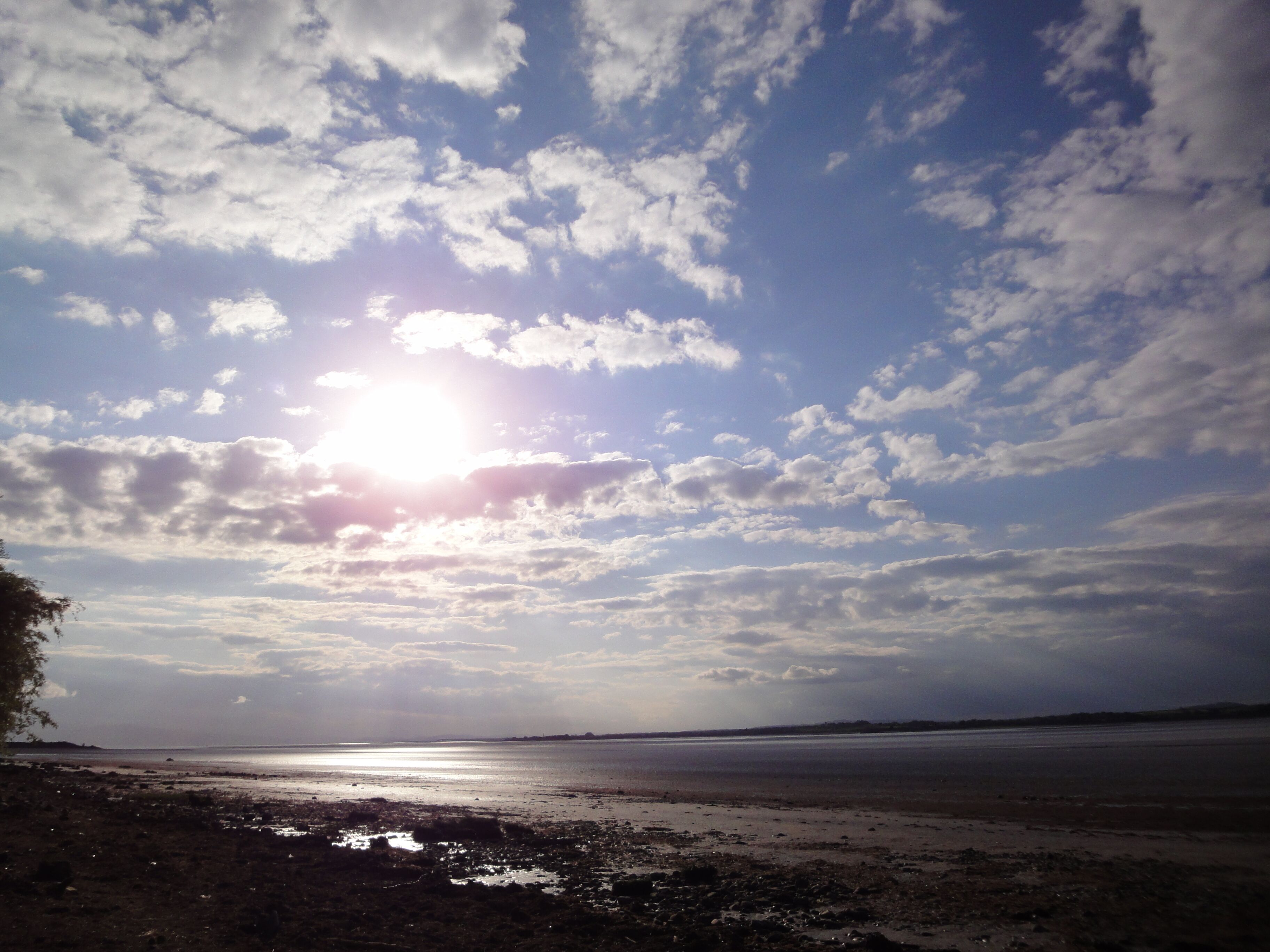 A beach with a clear sky and the sun shining through scattered clouds.
