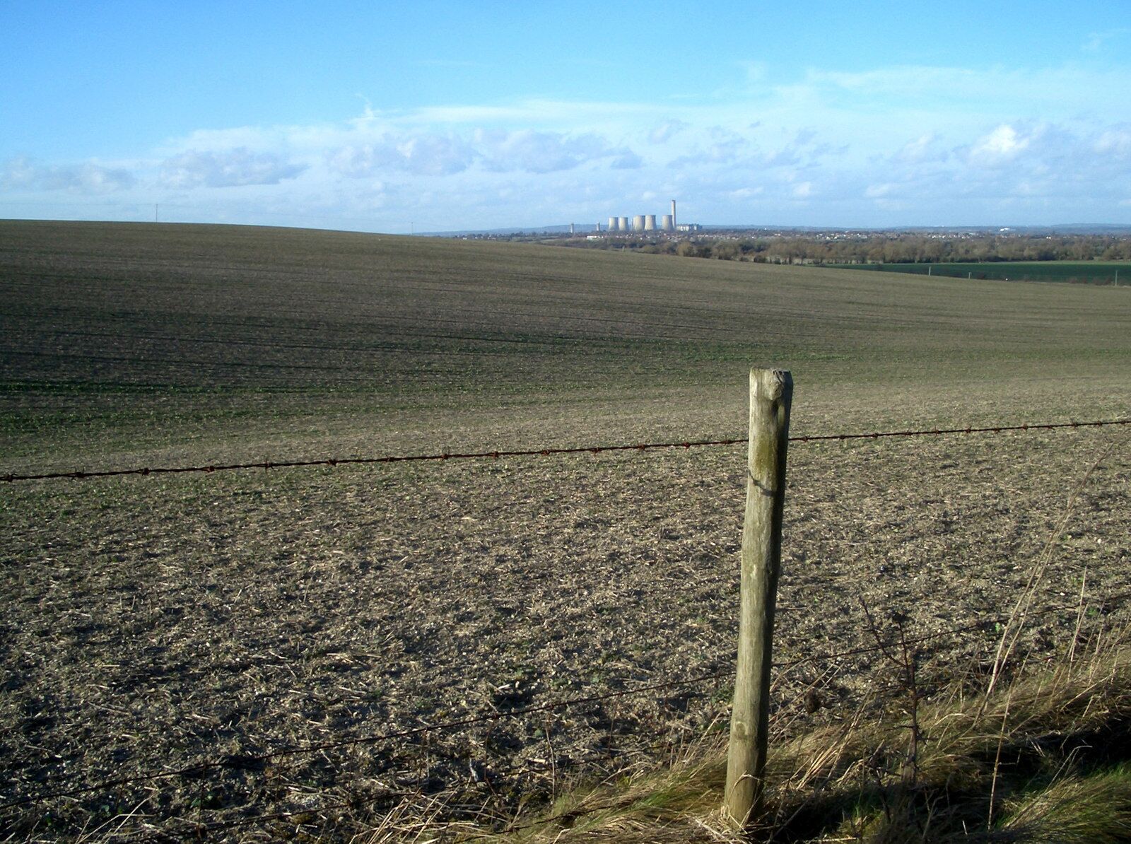 A vast, flat agricultural field with a barbed wire fence in the foreground and industrial buildings in the distance.