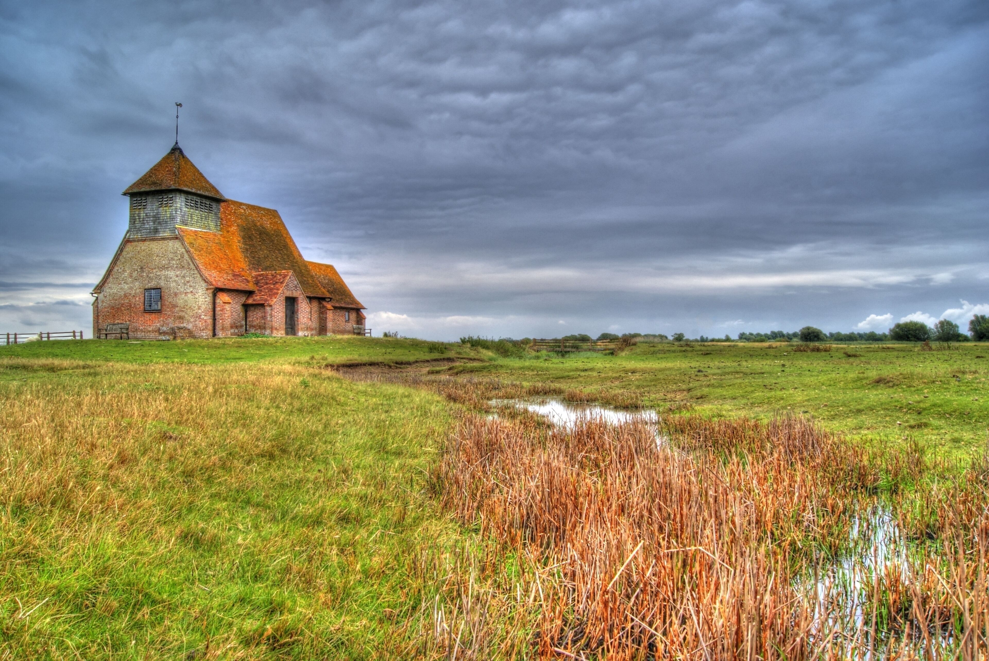 A brick church with a pointed roof and a tower, surrounded by a grassy field and a small pond.