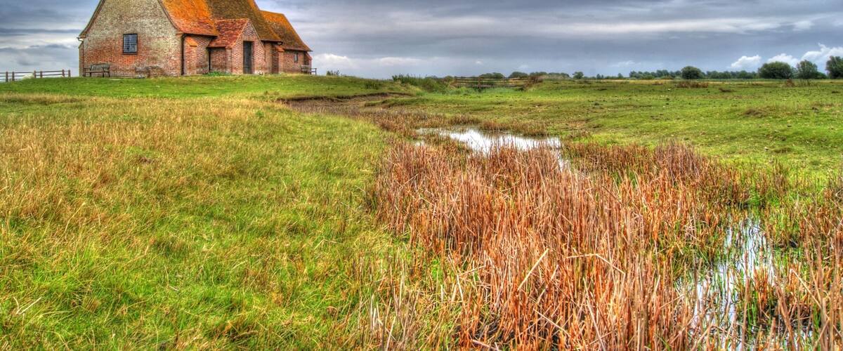 A brick church with a pointed roof and a tower, surrounded by a grassy field and a small pond.