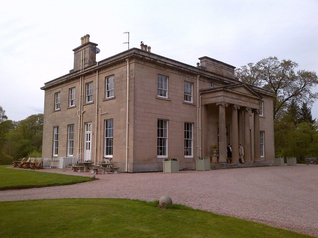 A large, historic building with a symmetrical facade, columns, and a gravel driveway.