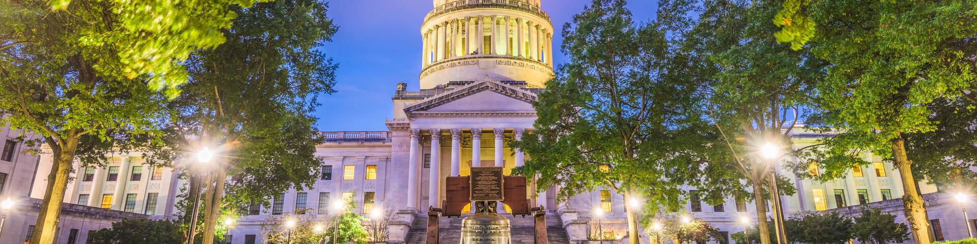 The West Virginia State Capitol building at dusk with a large bell in front.