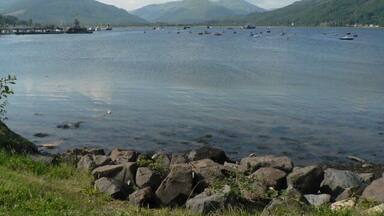 A calm lake with boats, rocky shoreline, and mountains in the background.