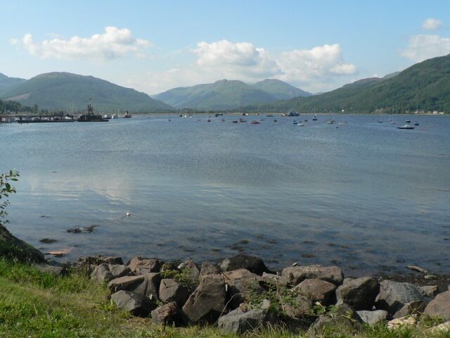 A calm lake with boats, rocky shoreline, and mountains in the background.