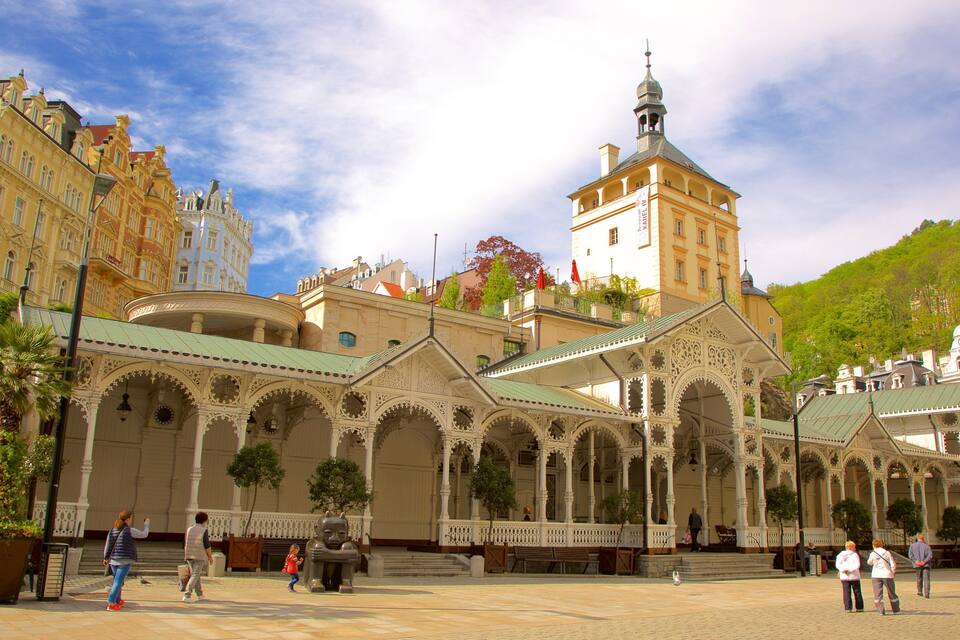 Um edifício histórico com arquitetura ornamentada, uma torre imponente e uma praça ao redor com pessoas e árvores.