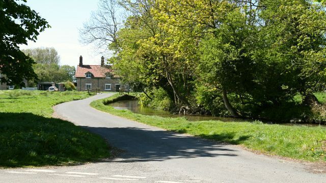 Off Malpas Road. The photograph is looking in a westerly direction along a short loop off Malpas Road (in the immediate foreground) toward Brooke Cottage (with red tiles roof - private property). The viewpoint is at the clockwise junction with Malpas Road. The eastern branch of Brompton Beck flows right to left (westerly) in the picture until it reaches the bridge (where the road rises slightly against the level of the water). At this point it is joined by the western branch of the beck coming from 428681 and turns south under the bridge. For a more upstream (left) photograph of the eastern branch of the beck, click here 428970. For a more downstream (ahead) photograph showing the beck, click here 428558. Behind the viewpoint is Brompton Butts - an area of grassland and trees. For a more anticlockwise (ahead) photograph of the loop, click here 428558. For a north-easterly (right) photograph of Malpas Road, click here 428955. For a south-westerly (left) photograph of Malpas Road, click here 427856.