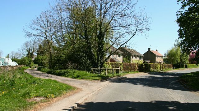 A rural road with houses on one side and a grassy area with trees on the other.